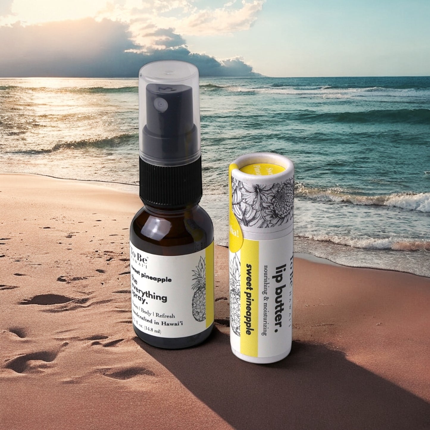 A lip butter and an everything spray products on a sandy beach with ocean waves in the background