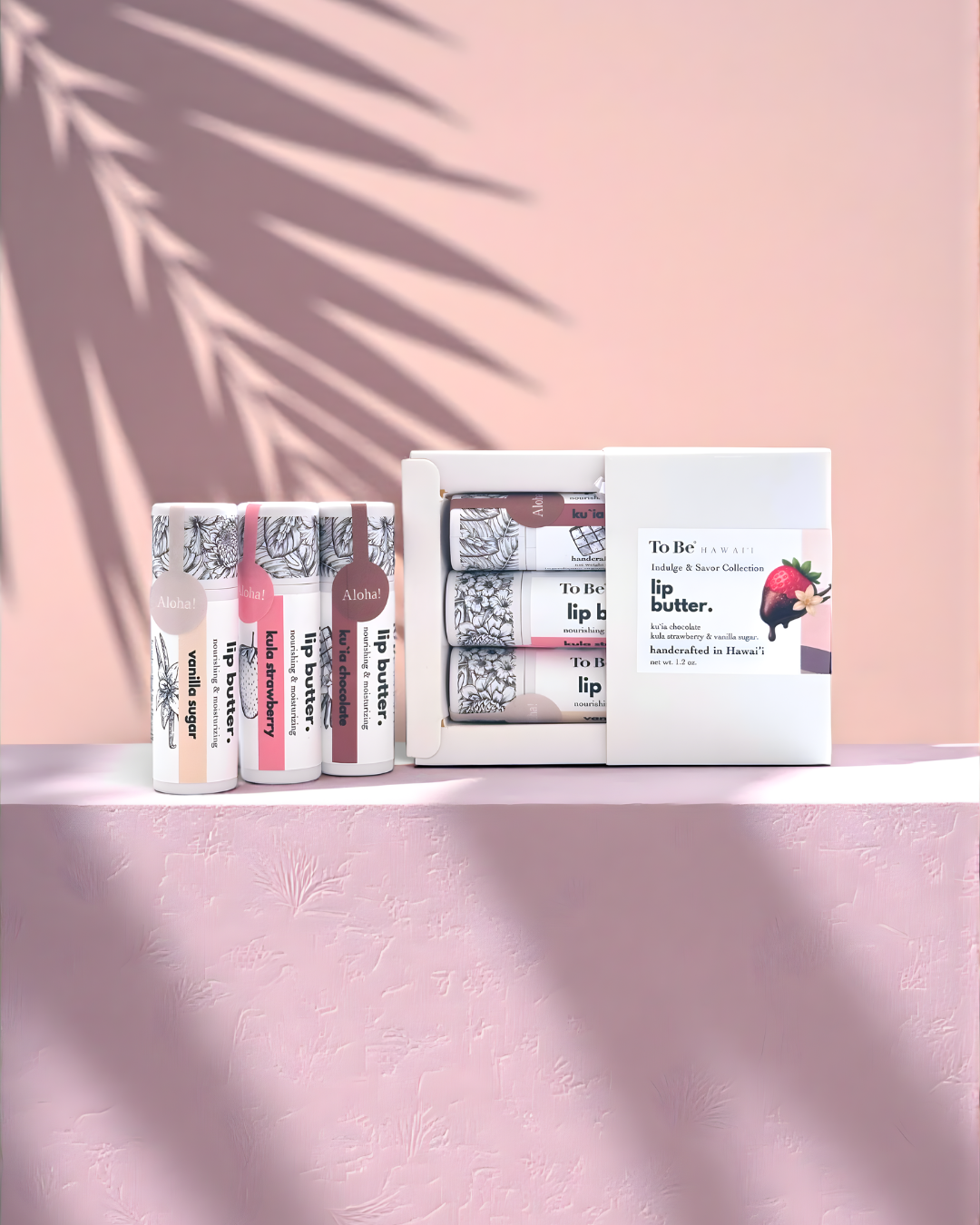 3 lip butters in a gift set for valentines day. Vanilla Sugar, Kula Strawberry and Kuia Chocolate sit on a pin surface with soft palm shadows in the background.