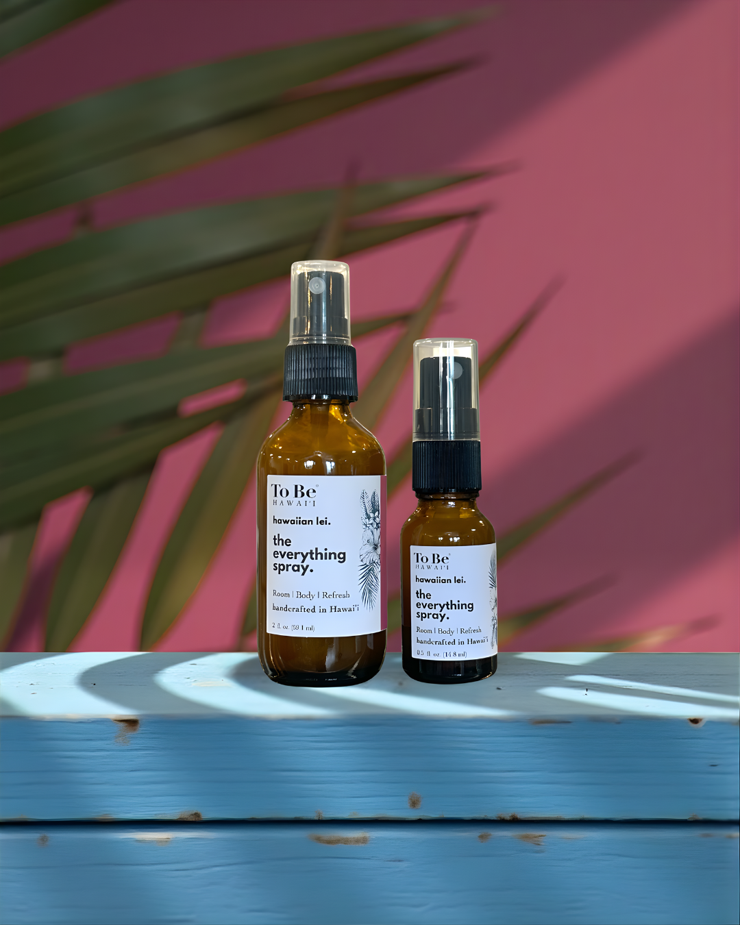 A large  bottle and a small bottle of the  To Be brand everything spray hawaiian lei with labels on a blue wood surface with a red and green leafy background.