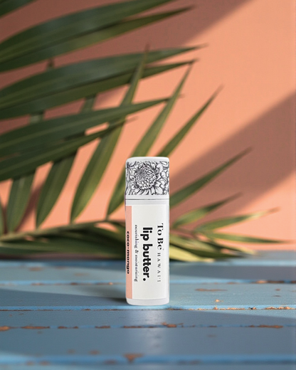 Coco-Mango Lip butter in a cardboard push up recyclable tube on a blue surface with green leaves in the background