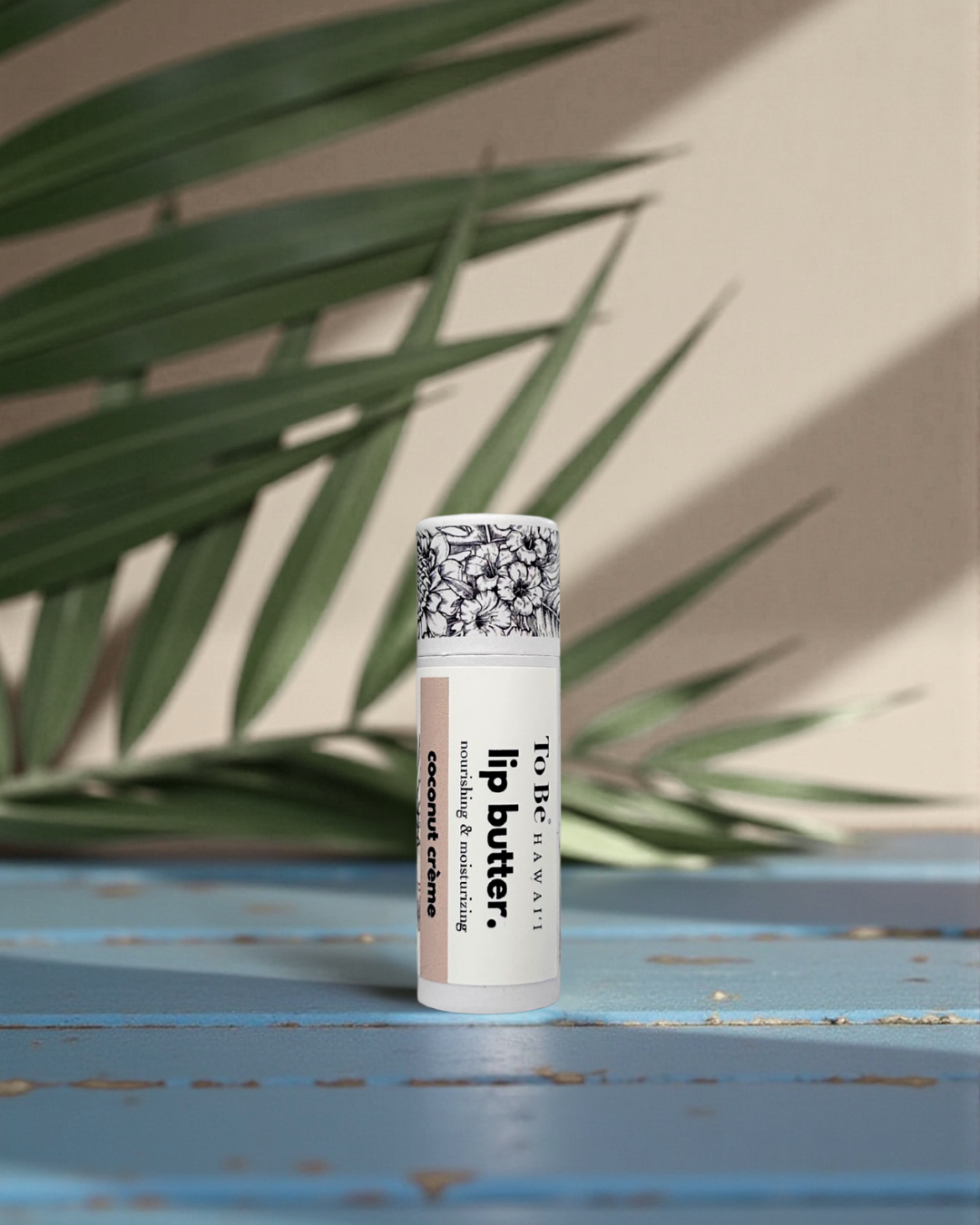 Coconut Crème Lip butter in a cardboard push up recyclable tube on a blue surface with green leaves in the background