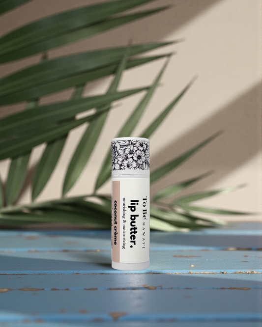 Coconut Crème Lip butter in a cardboard push up recyclable tube on a blue surface with green leaves in the background