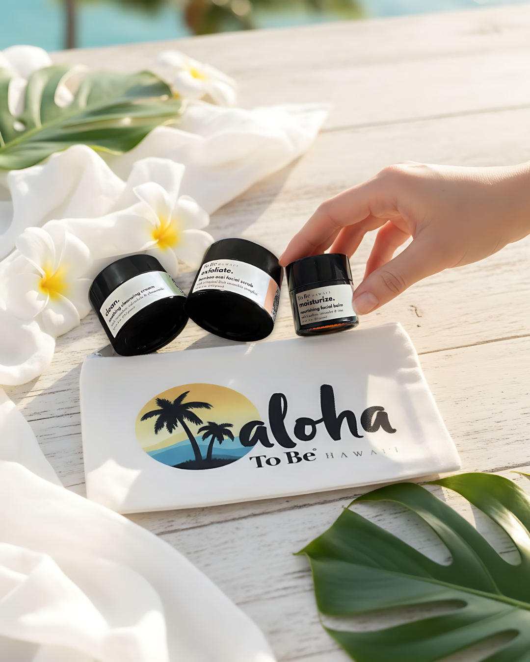 The To Be skin care discovery set with 3 glass jars with black lids on a white table featuring 'Aloha To Be' branded zip canvas bag, surrounded by white flowers and green leaves.