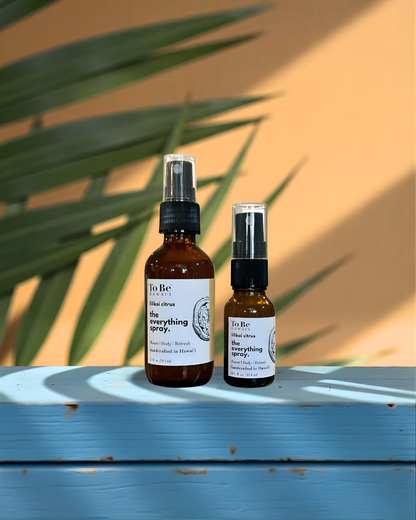 Two bottles of 'To Be' brand lilikoi citrus everything spray on a blue surface with a orange wall and green leaves in the background.