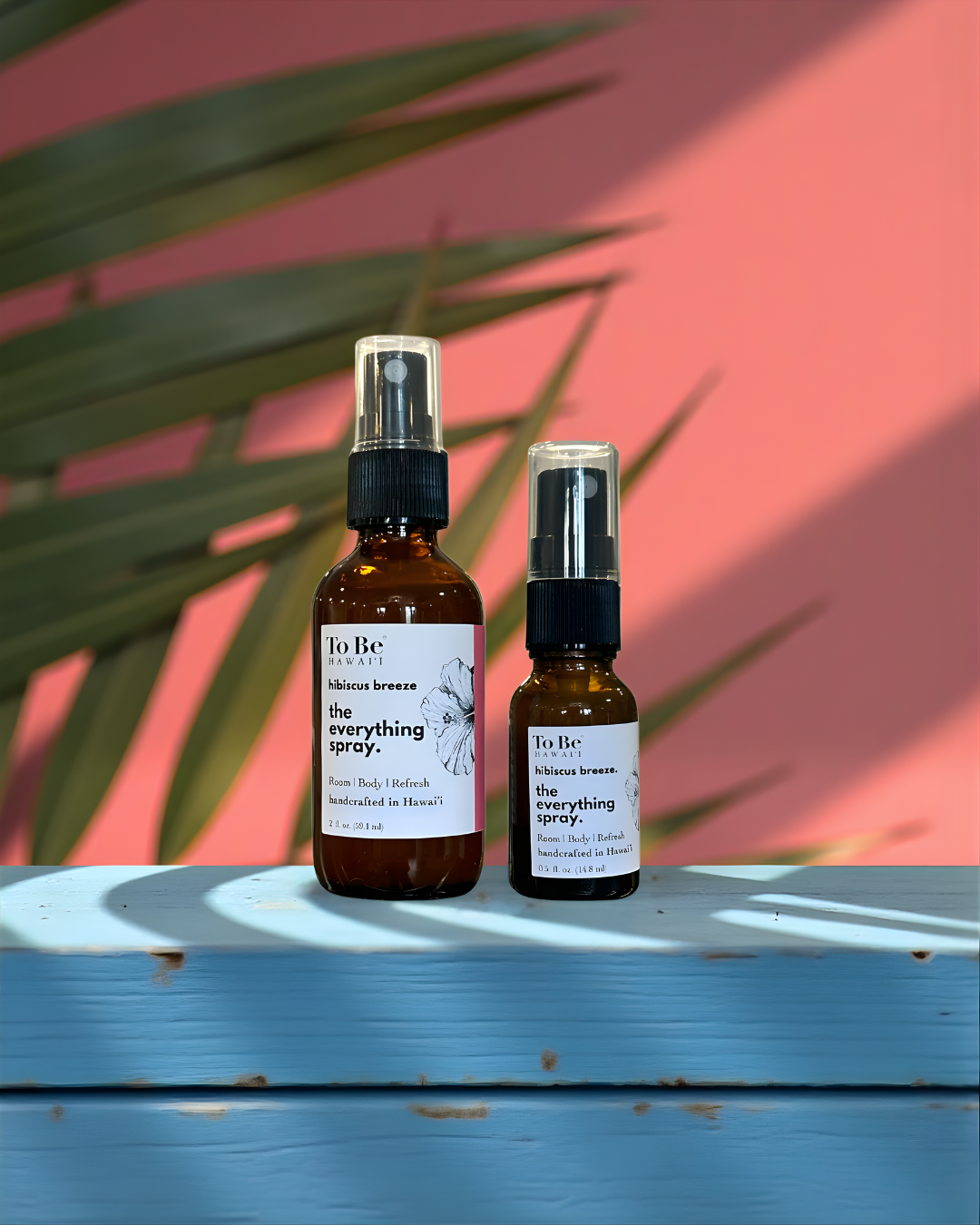 A large  bottle and a small bottle of the  To Be brand everything spray hibiscus breeze with labels on a blue wood surface with a red and green leafy background.