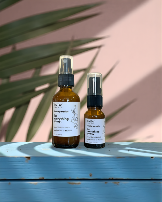 A large  bottle and a small bottle of the  To Be brand everything spray pikake paradisewith labels on a blue wood surface with a peach and green leafy background.