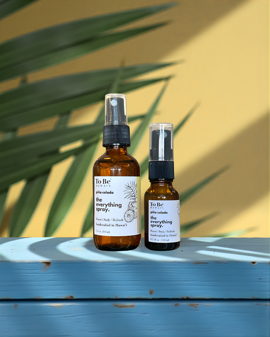 A large  bottle and a small bottle of the  To Be brand everything spray pina colada with labels on a blue wood surface with a yellow and green leafy background.