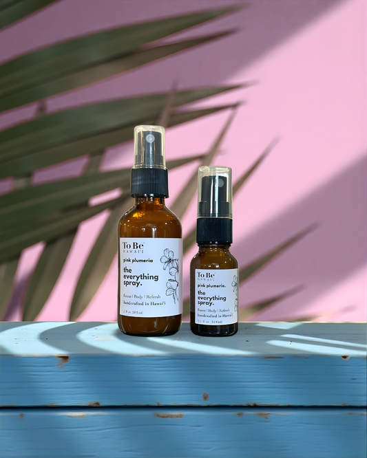 A large  bottle and a small bottle of the  To Be brand everything spray pink plumeria with labels on a blue wood surface with a pink and green leafy background.