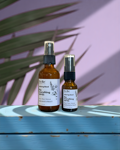 A large  bottle and a small bottle of the  To Be brand everything spray relax with labels on a blue wood surface with a lavender and green leafy background.