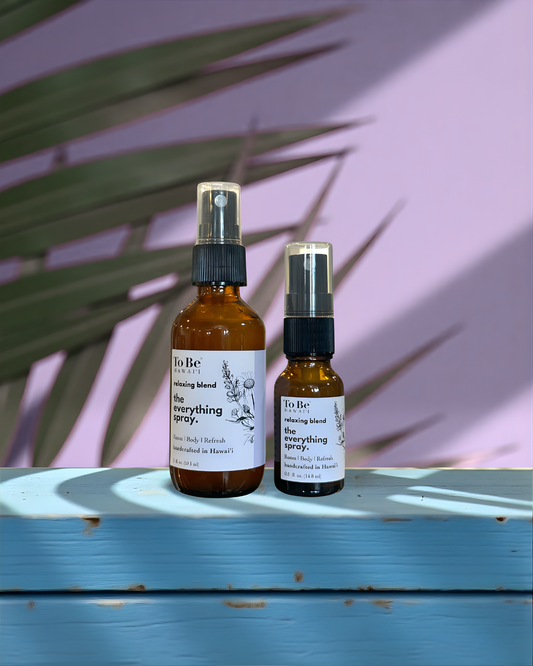A large  bottle and a small bottle of the  To Be brand everything spray relax with labels on a blue wood surface with a lavender and green leafy background.