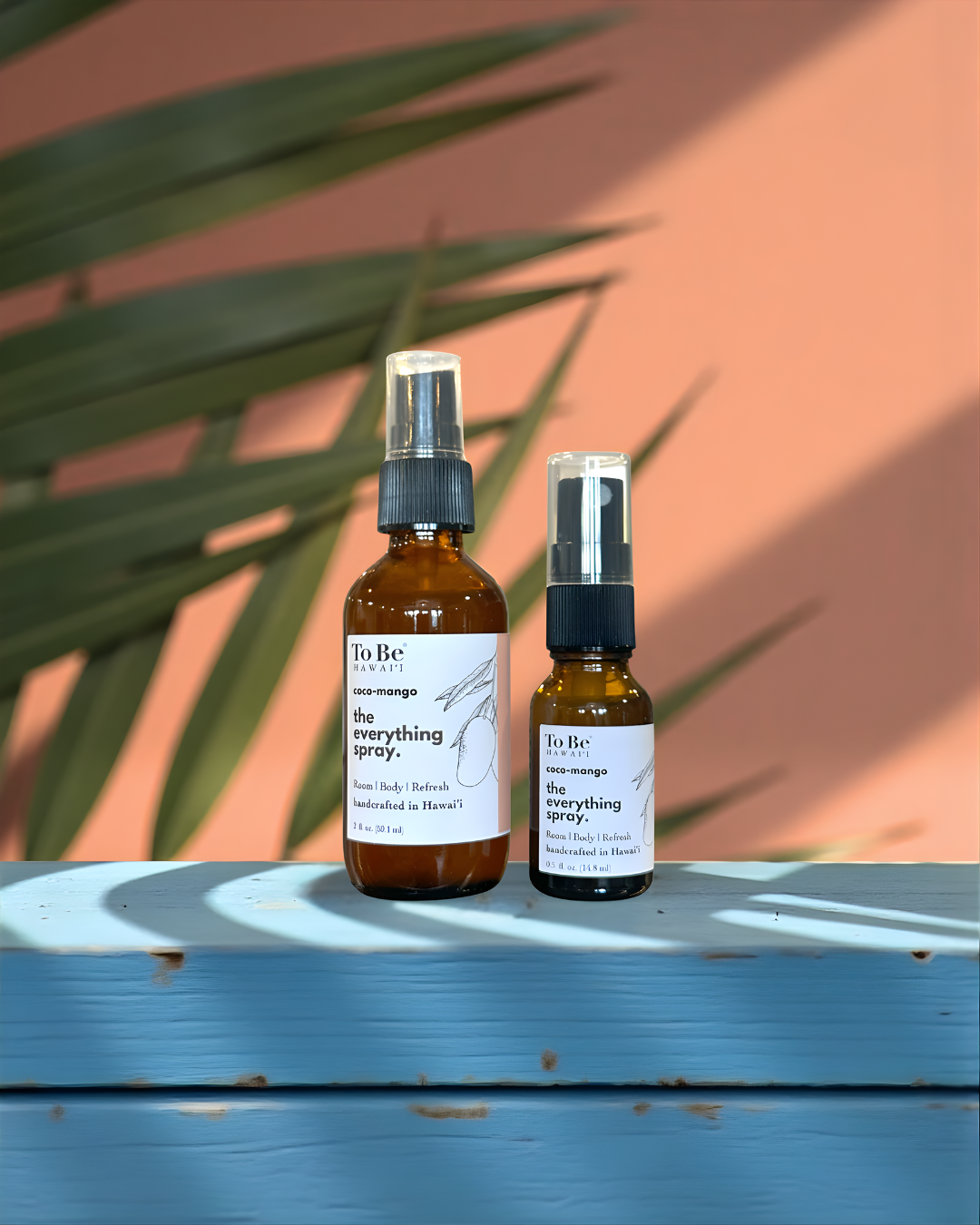 Two bottles of 'To Be' brand Coco-mango everything spray on a blue surface with a orange wall and green leaves in the background.