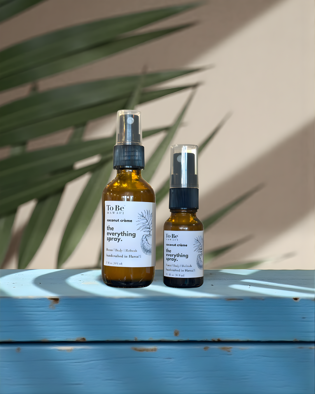 Two bottles of 'To Be' brand coconut creme everything spray on a blue surface with a beige wall and green leaves in the background.