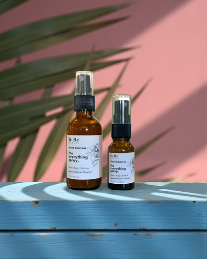 A large  bottle and a small bottle of the everything spray Island tuberose  with labels on a blue wood surface with a pink and green leafy background.