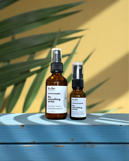 Two bottles of 'To Be' brand pineapple everything spray on a blue surface with a yellow wall and green leaves in the background.