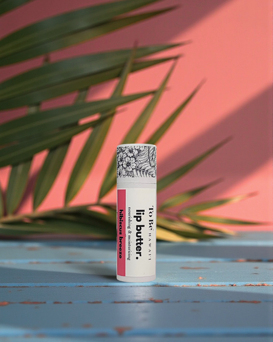 Hibiscus Breeze Lip butter in a cardboard push up recyclable tube on a blue surface with green leaves in the background