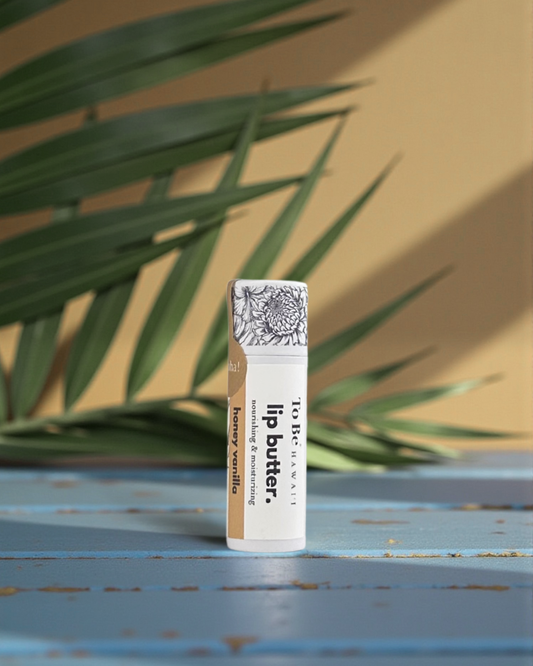 Honey Vanilla  Lip butter in a cardboard push up recyclable tube on a blue surface with green leaves in the background