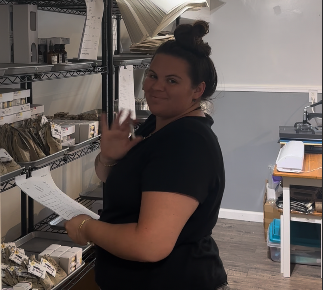 Person in a black shirt standing in a storage or office area with shelves and a desk.