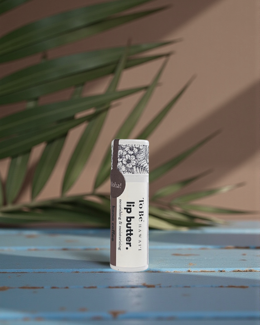 Kona Coffee Lip butter in a cardboard push up recyclable tube on a blue surface with green leaves in the background