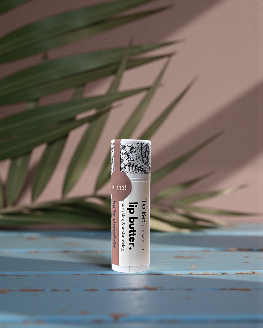 Ku`ia Chocolate Lip butter in a cardboard push up recyclable tube on a blue surface with green leaves in the background