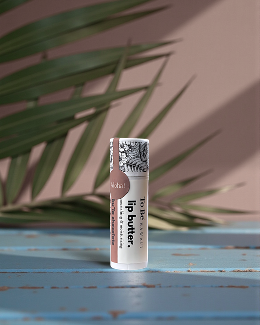 Ku`ia Chocolate Lip butter in a cardboard push up recyclable tube on a blue surface with green leaves in the background