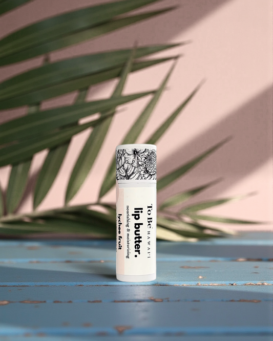 Lychee Fruit  Lip butter in a cardboard push up recyclable tube on a blue surface with green leaves in the background
