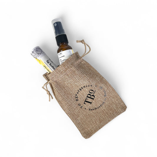 a small everything spray bottles and a lip butter in a burlap drawstring bag with 'TBO' branding.