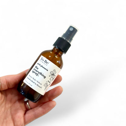 A hand holding a brown amber glass bottle with a white label that reads 'The Everything Spray Pink Plumeria', equipped with a fine mister for spraying.