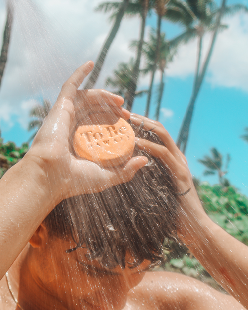Person holding a shampoo bar  with 'To Be' text under a shower, palm trees in the background