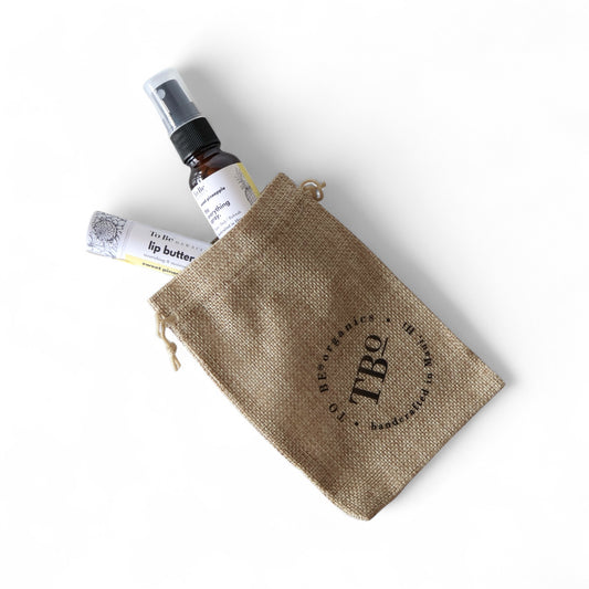a small everything spray bottles and a lip butter in a burlap drawstring bag with 'TBO' branding.