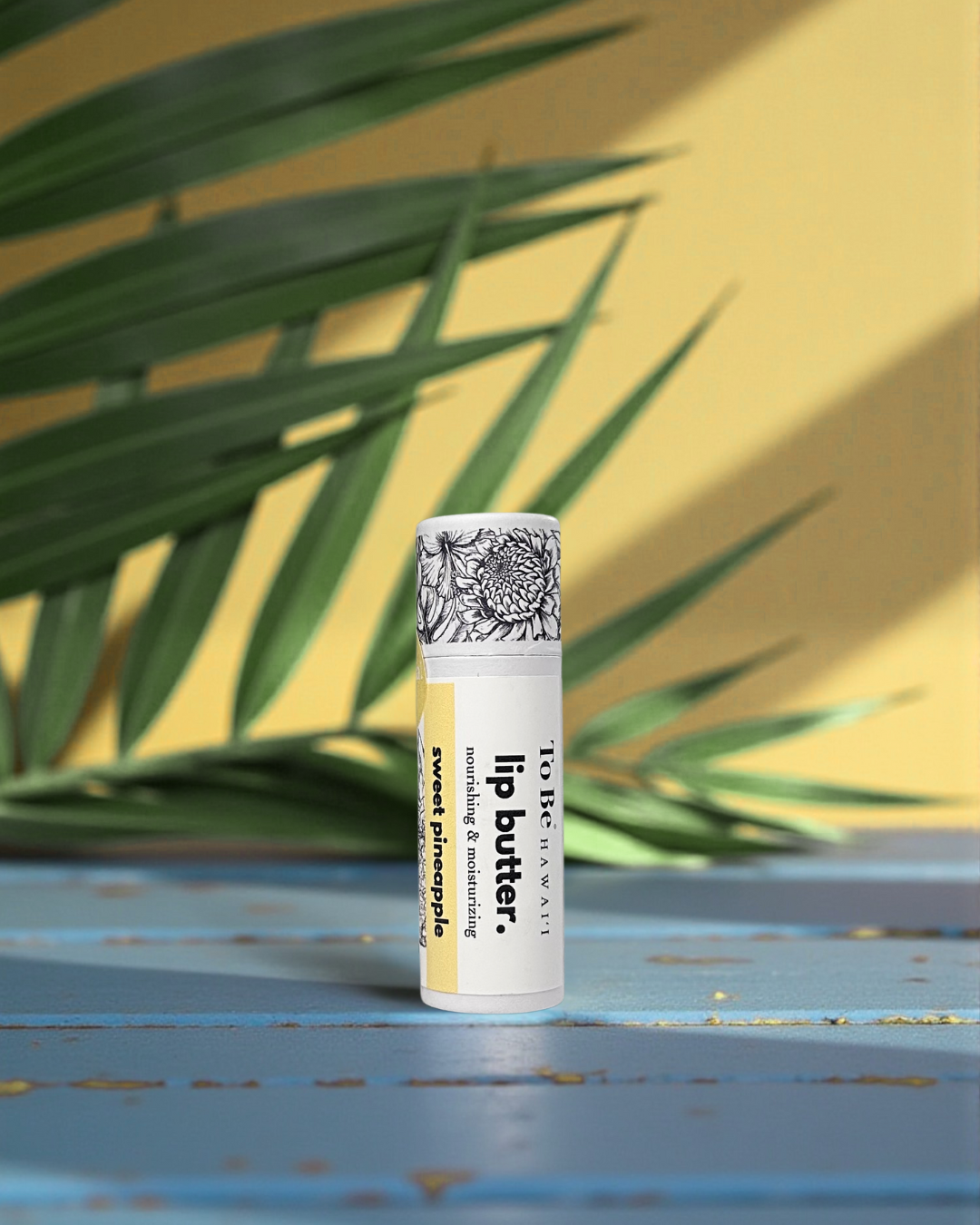 Sweet Pineapple Lip butter on a blue surface with green leaves in the background
