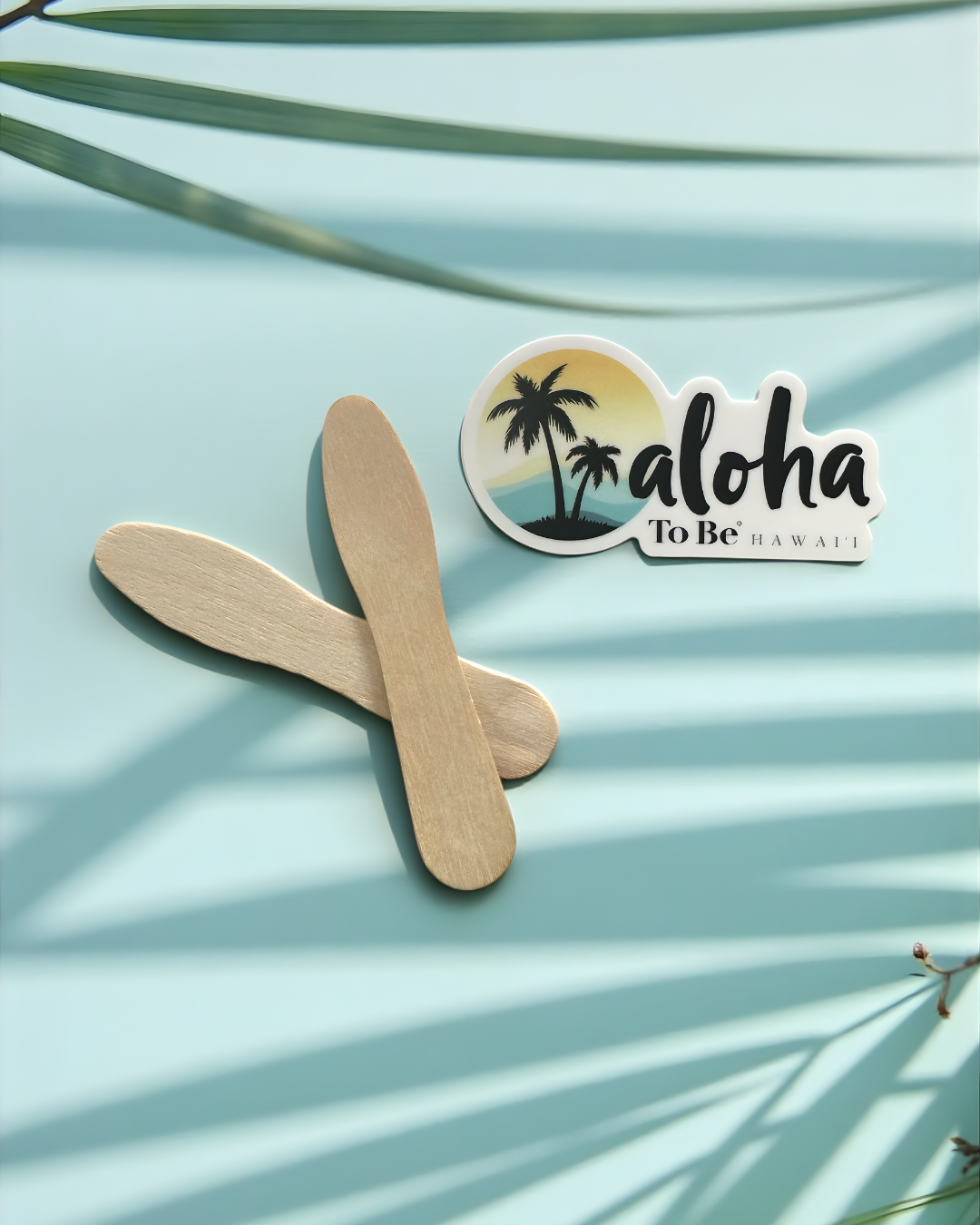 Two wooden sticks on a light blue background with a ' aloha To Be Hawaii' sticker.