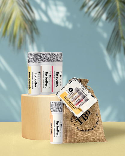 Four lip butters, sweet pineapple, coconut creme, hibiscus breeze, and lilikoi citrus with burlap drawstring bag on a yellow surface.