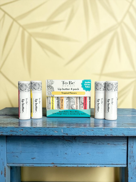 A set of 4 lip butter tubes and one box of lip butter arranged on a distressed blue wooden table against a pale yellow wall with a leaf pattern. The brand name 'To Be’ is visible on the packaging.