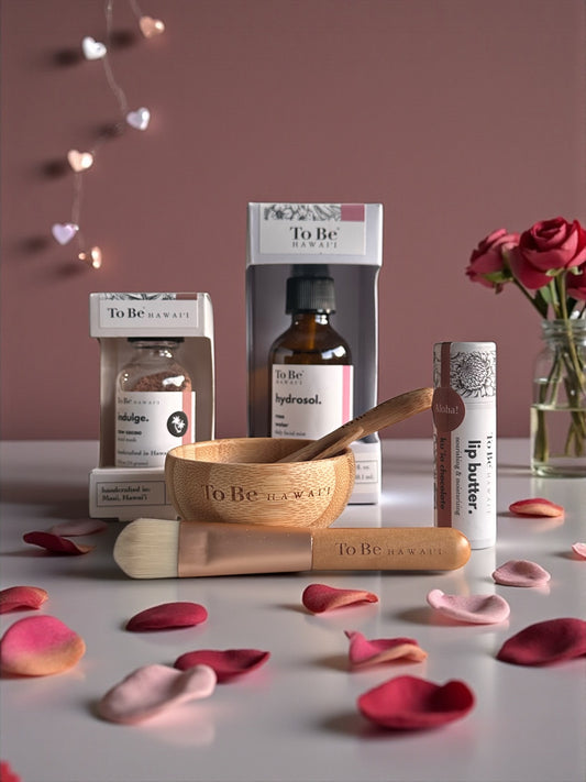 A rose hydrosol, chocolate mask, chocolate lip butter and bamboo bowl and brush surrounded by rose petals.