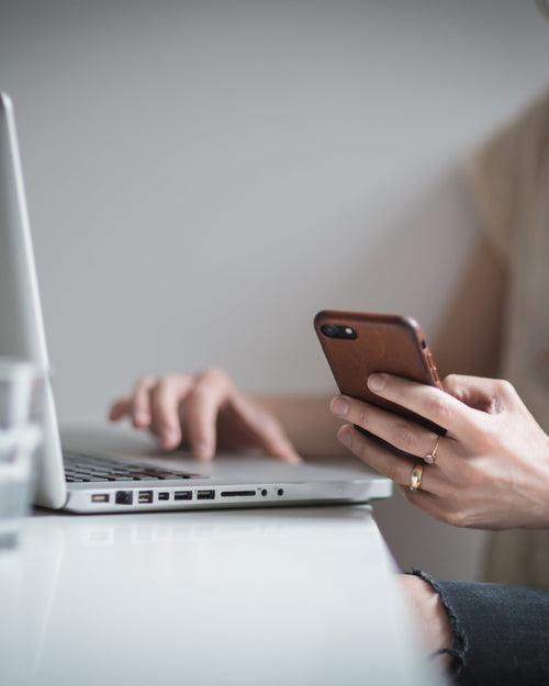 Woman sitting at desk checking her laptop and checking her iphone.